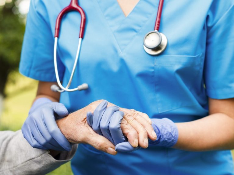 A compassionate healthcare professional in blue scrubs and gloves holds the hand of an elderly patient, symbolizing personalized support in Ambetter Approved Argent Care.