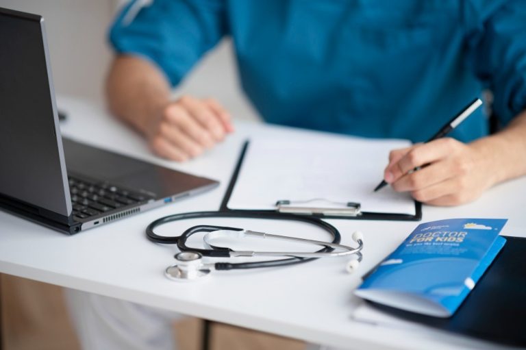Medical professional in clinical setting writing notes beside laptop and pediatric health pamphlet at Walk-In Clinic Houston.
