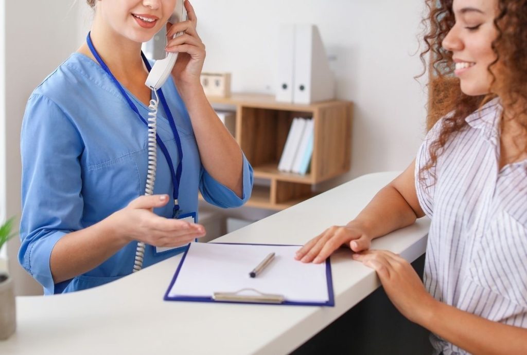 Receptionist in blue scrubs speaking on the phone at a Walk-In Clinic Houston front desk, assisting a patient with check-in and appointment coordination.