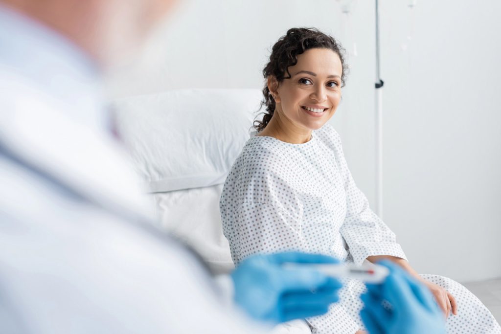 Gloved hands hold a tool as a patient in a gown smiles in a bed
