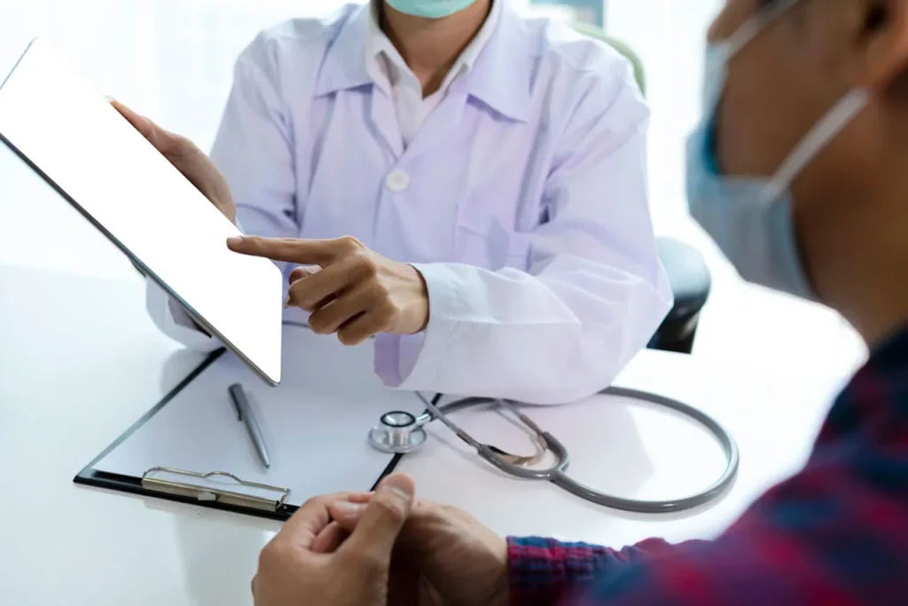 A masked doctor uses a tablet to review test results and discuss health with a patient during an annual physical exam.
