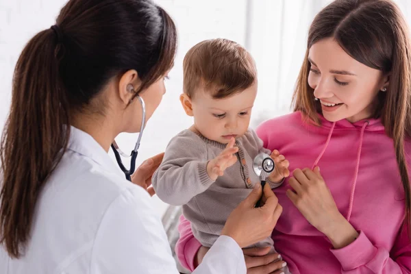 Female primary care pediatrician using a stethoscope to examine a baby held by his mother.